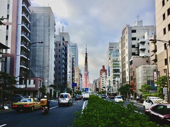 A two way street in Tokyo on a cloudy day, separated by a hedge. It is bordered by tall apartment buildings and on the horizon stands a white a red metallic tower. A scooter, a yellow taxi and other cars are on each side of the street.